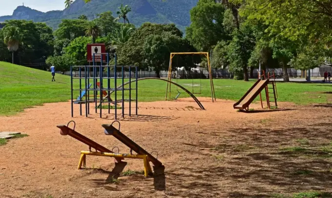 Parque com equipamentos públicos de lazer, incluindo brinquedos e estruturas de ginástica, em área verde com árvores e montanhas ao fundo