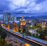 Medellín, Colômbia, destacando a arquitetura moderna, prédios iluminados, uma estação de metrô em movimento e montanhas ao fundo.