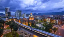 Medellín, Colômbia, destacando a arquitetura moderna, prédios iluminados, uma estação de metrô em movimento e montanhas ao fundo.