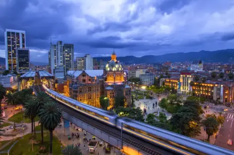 Medellín, Colômbia, destacando a arquitetura moderna, prédios iluminados, uma estação de metrô em movimento e montanhas ao fundo.