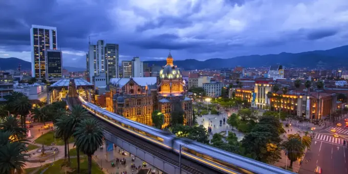 Medellín, Colômbia, destacando a arquitetura moderna, prédios iluminados, uma estação de metrô em movimento e montanhas ao fundo.