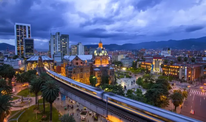 Medellín, Colômbia, destacando a arquitetura moderna, prédios iluminados, uma estação de metrô em movimento e montanhas ao fundo.