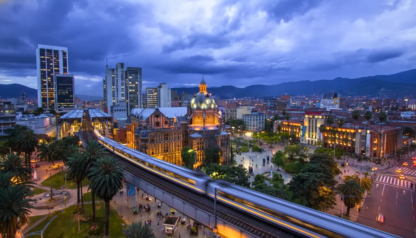 Medellín, Colômbia, destacando a arquitetura moderna, prédios iluminados, uma estação de metrô em movimento e montanhas ao fundo.