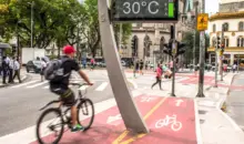 Ciclovia ativa em uma rua movimentada de uma cidade, com ciclistas, pedestres, sinalizações e clima de calor de 30°C, destacando a pegada ambiental da IA na mobilidade sustentável.