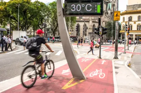 Ciclovia ativa em uma rua movimentada de uma cidade, com ciclistas, pedestres, sinalizações e clima de calor de 30°C, destacando a pegada ambiental da IA na mobilidade sustentável.