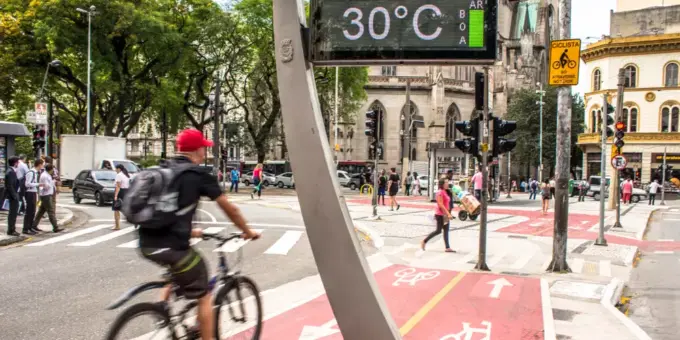Ciclovia ativa em uma rua movimentada de uma cidade, com ciclistas, pedestres, sinalizações e clima de calor de 30°C, destacando a pegada ambiental da IA na mobilidade sustentável.