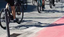 Grupo de ciclistas pedalando em ciclovia pavimentada, promovendo transporte sustentável e saúde em área urbana com sombra.