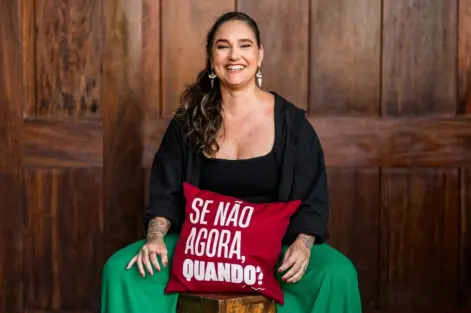 Lídice Berman sentada em um banco de madeira, apoiada em um travesseiro vermelho