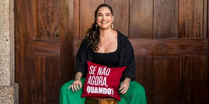 Lídice Berman sentada em um banco de madeira, apoiada em um travesseiro vermelho
