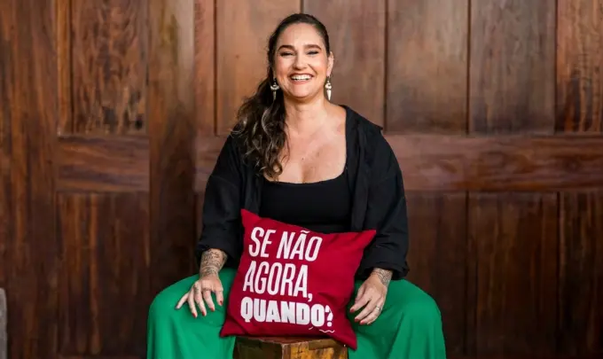 Lídice Berman sentada em um banco de madeira, apoiada em um travesseiro vermelho