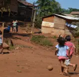 Ocupação tradicional do povo indígena Guarani-Mbya, no Jaraguá, região oeste. Crianças brincando ao ar livre em uma área rural com casas de madeira e vegetação ao fundo, representando uma comunidade carente.