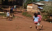 Ocupação tradicional do povo indígena Guarani-Mbya, no Jaraguá, região oeste. Crianças brincando ao ar livre em uma área rural com casas de madeira e vegetação ao fundo, representando uma comunidade carente.