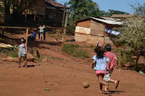Ocupação tradicional do povo indígena Guarani-Mbya, no Jaraguá, região oeste. Crianças brincando ao ar livre em uma área rural com casas de madeira e vegetação ao fundo, representando uma comunidade carente.