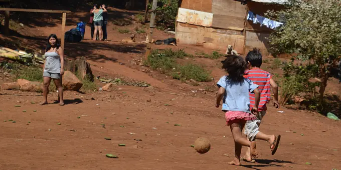 Ocupação tradicional do povo indígena Guarani-Mbya, no Jaraguá, região oeste. Crianças brincando ao ar livre em uma área rural com casas de madeira e vegetação ao fundo, representando uma comunidade carente.