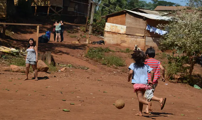 Ocupação tradicional do povo indígena Guarani-Mbya, no Jaraguá, região oeste. Crianças brincando ao ar livre em uma área rural com casas de madeira e vegetação ao fundo, representando uma comunidade carente.