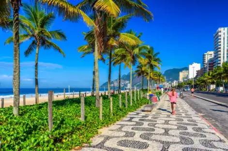 Dia ensolarado na orla da praia de Ipanema, com uma pedestre passando. A foto ilustra a matéria sobre a restauração da restinga em Ipanema.
