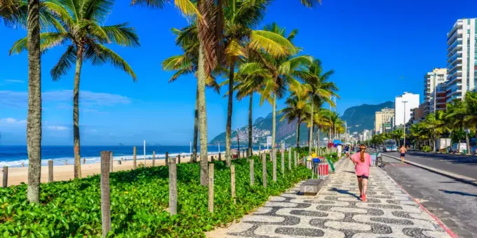 Dia ensolarado na orla da praia de Ipanema, com uma pedestre passando. A foto ilustra a matéria sobre a restauração da restinga em Ipanema.