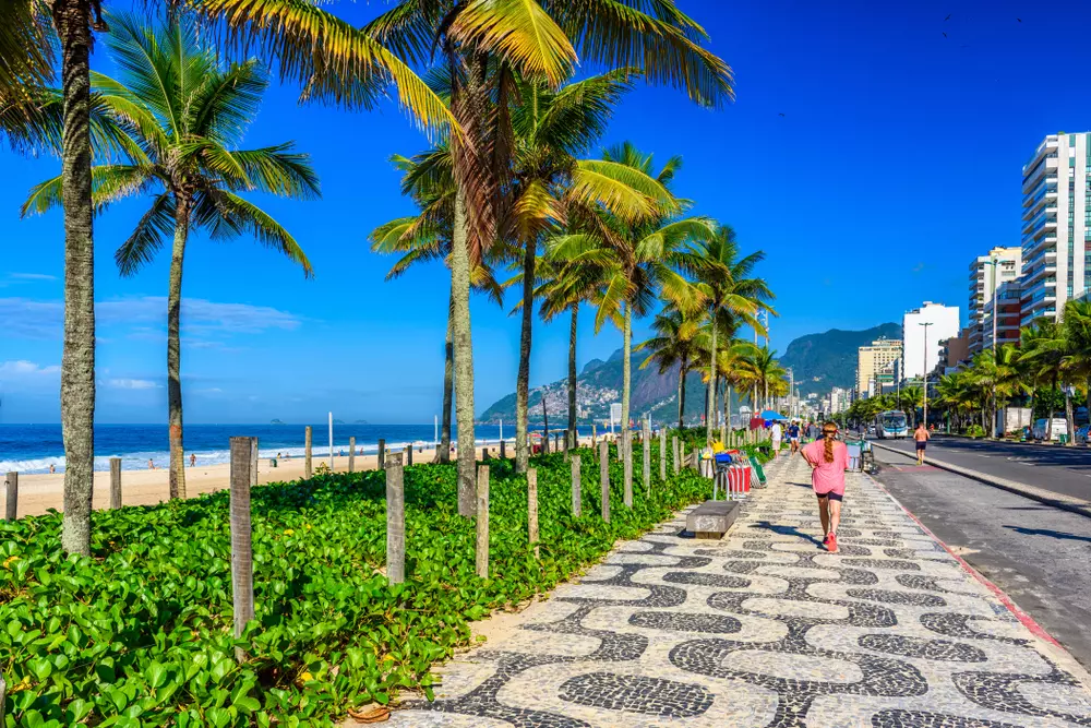Dia ensolarado na orla da praia de Ipanema, com uma pedestre passando. A foto ilustra a matéria sobre a restauração da restinga em Ipanema.