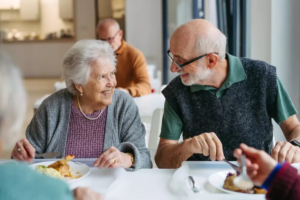 Casal de idosos sorrindo e desfrutando de uma refeição juntos em um ambiente acolhedor, refletindo momentos de felicidade e convivência na terceira idade.